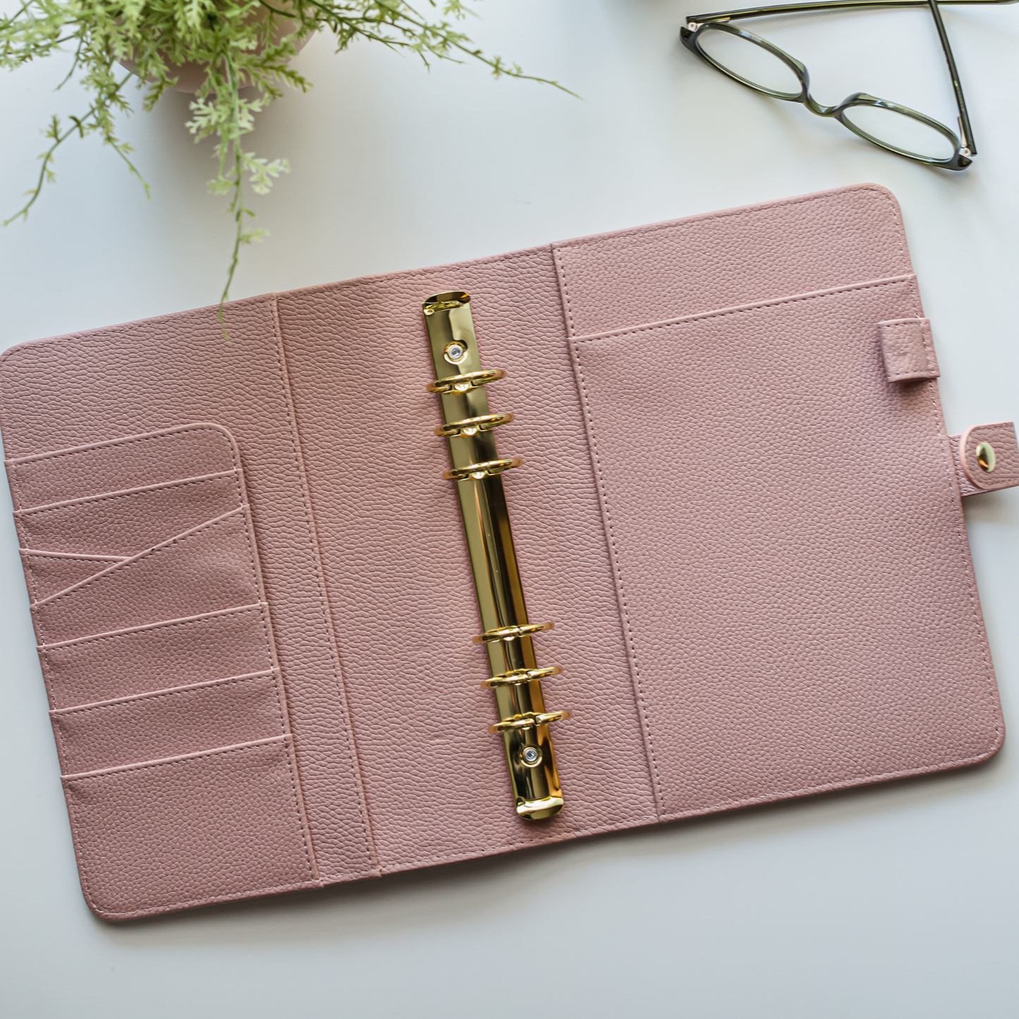 Pink planner with gold ring binder on a white surface with a plant and glasses in the background