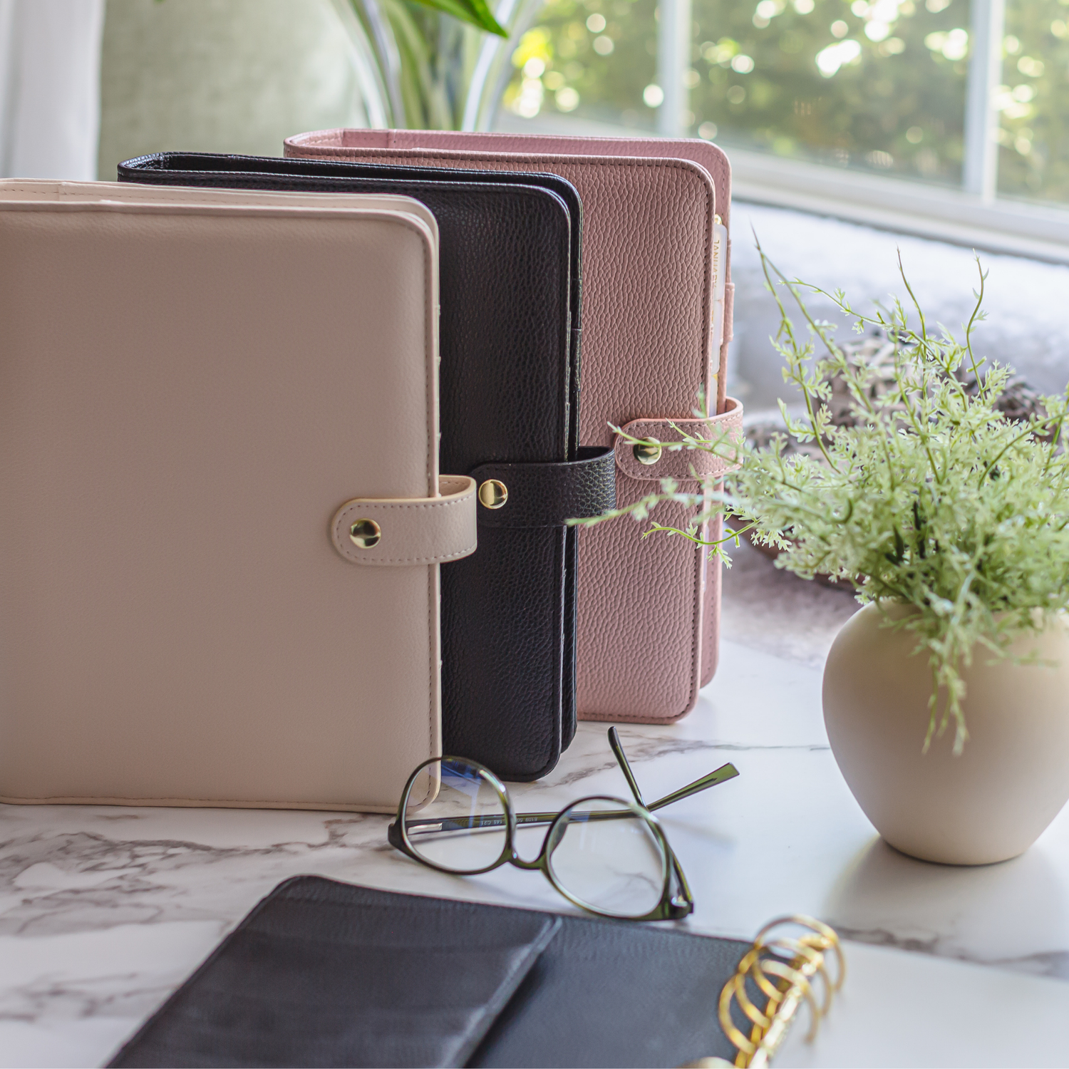 Three planner covers in beige, black, and pink on a marble surface with glasses and a plant.
